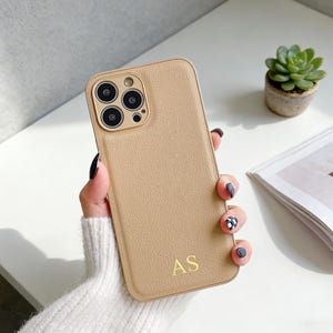 Puede incluir: Funda de teléfono de cuero color canela con las iniciales doradas "AS". La funda tiene una superficie texturizada y un recorte para la cámara. Una pequeña planta suculenta en maceta y una revista están en el fondo.