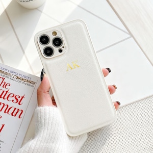 May include: A white phone case with gold-colored initials "AK" is held in a person's hand. The case has a textured surface and a camera module with three lenses. A coffee cup and a book are in the background.