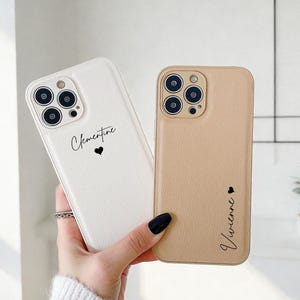 Puede incluir: Dos fundas de teléfono, una blanca y otra beige, se sostienen en una mano. La funda blanca tiene el nombre "Clementine" y un corazón, mientras que la funda beige tiene "Vivienne" y un corazón. Ambas fundas tienen un acabado texturizado.