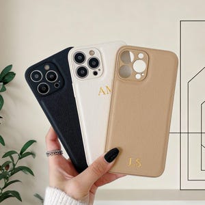 Puede incluir: Se muestran tres fundas de teléfono personalizadas en negro, blanco y beige. La funda blanca tiene las iniciales "AM" en dorado, y la beige "LS". Las fundas tienen un aspecto texturizado similar al cuero y están diseñadas para un smartphone.