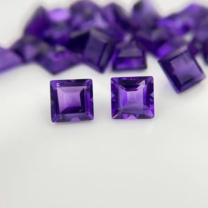 Könnte beinhalten: Zwei quadratisch geschliffene, facettierte Amethyst-Edelsteine ​​stehen im Fokus und zeigen ihre tiefviolette Farbe und inneren Facetten. Andere Amethyststeine ​​sind im Hintergrund unscharf. Die Edelsteine ​​sind vor einem weißen Hintergrund platziert.