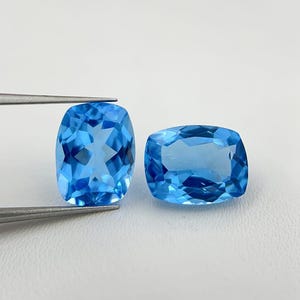 Könnte beinhalten: Zwei hellblaue Edelsteine mit vielen Facetten. Einer wird von einer Pinzette gehalten, der andere liegt daneben. Die Edelsteine haben einen rechteckigen Kissenschliff und reflektieren das Licht. Der Hintergrund ist eine schlichte weiße Oberfläche.