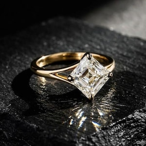 Könnte beinhalten: Ein Goldring mit einem einzelnen, im Prinzessschliff facettierten, klaren Edelstein in einer Vier-Krappen-Fassung. Die Ringschiene hat ein geteiltes Design, das sich zur Mitte hin verjüngt, mit polierter Metalloberfläche und quadratischem Stein.