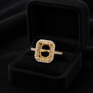 May include: A gold ring featuring a rectangular halo setting designed for a center stone. The band and the rectangular halo are encrusted with small, clear pavé-set stones. The ring is presented in a black velvet jewelry box.