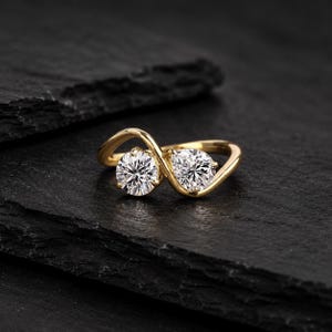 Op de afbeelding: Een gouden bypass-ring met twee ronde, briljant geslepen heldere stenen die naast elkaar zijn geplaatst. De ringband heeft een gladde, gepolijste afwerking die om elke steen heen buigt, wat zorgt voor een open en vloeiend ontwerp.