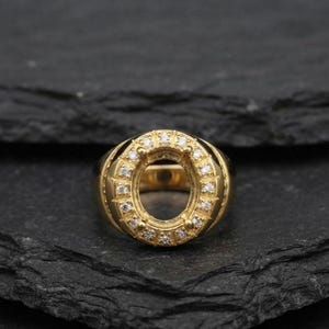 May include: A gold ring featuring an oval-shaped center setting designed to hold a gemstone. The band and the oval frame are crafted from polished yellow gold, with the oval frame accented by a series of small, clear, round-cut white stones.