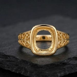 May include: A gold ring setting featuring a central cushion-shaped bezel mount designed to hold a gemstone. The band displays intricate openwork filigree patterns along the sides, showcasing a polished metallic finish and a hollow center.