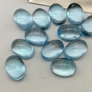Könnte beinhalten: Eine Ansammlung von elf glatten, oval geformten hellblauen Glaskabochons mit polierter Oberfläche. Jedes Stück ist durchscheinend und reflektiert das Licht, wobei das gesamte Set einen einheitlichen hellblauen Farbton aufweist, angeordnet neben einer Metallschaufel.