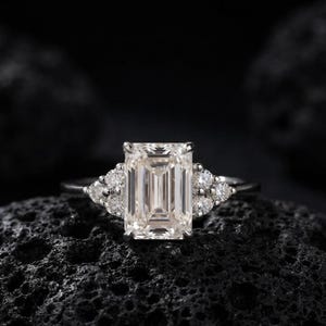 Könnte beinhalten: Ein silberfarbener Ring mit einem großen, rechteckigen Mittelstein im Smaragdschliff. Der Mittelstein ist auf jeder Seite von einer Gruppe aus drei kleineren, rund geschliffenen Steinen flankiert. Die Ringschiene ist dünn und metallisch.