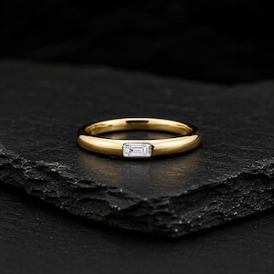 Könnte beinhalten: Ein polierter Ring aus Gelbgold mit einem einzelnen, horizontal gefassten, rechteckigen, klaren Edelstein im Baguetteschliff in der Mitte. Der Ring hat ein glattes, abgerundetes Profil und eine hochglänzende metallische Oberfläche.