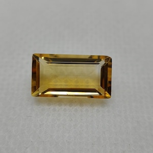 Könnte beinhalten: Ein rechteckiger, facettierter Citrin-Edelstein mit einem transparenten, goldgelben Farbton. Der Stein weist einen Treppenschliff mit scharfen, sauberen Kanten und eine polierte Oberfläche auf, die das Licht reflektiert und seine klare, warme Farbe zur Geltung bringt.