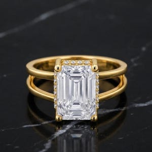 Peut inclure: Une bague en or avec une grande pierre transparente rectangulaire taille émeraude en son centre. La pierre est entourée d'un délicat rebord de minuscules pierres blanches étincelantes. L'anneau est divisé en deux bandes dorées polies.