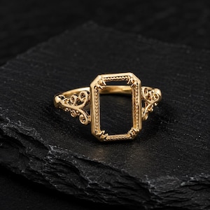 Könnte beinhalten: Ein goldfarbener Ring mit einer zentralen rechteckigen Fassung, die einen perlenartigen Rand und vier kleine Krappen aufweist. Die Ringschiene ist auf beiden Seiten mit filigranen Schnörkelmustern verziert, die vom rechteckigen Rahmen ausgehen.