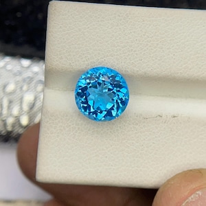 Könnte beinhalten: Ein runder, facettierter Edelstein in leuchtendem Hellblau. Der Stein ist im Brillantschliff gehalten und besitzt zahlreiche dreieckige und rautenförmige Facetten, die das Licht reflektieren und auf einem weißen, strukturierten Sockel funkeln.