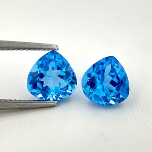 Könnte beinhalten: Zwei tropfenförmige, facettierte blaue Edelsteine. Einer wird von einer silbernen Pinzette gehalten. Die Edelsteine haben einen brillanten, facettenreichen Schliff, der das Licht reflektiert. Die Edelsteine sind leuchtend blau, vor weißem Hintergrund.