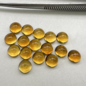 Könnte beinhalten: Eine Sammlung von runden, bernsteinfarbenen Edelsteinen. Die im Cabochon-Schliff geschliffenen Steine sind auf einer hellen Oberfläche angeordnet, mit einer silbernen Pinzette im Hintergrund. Die Edelsteine haben ein poliertes, glänzendes Aussehen.