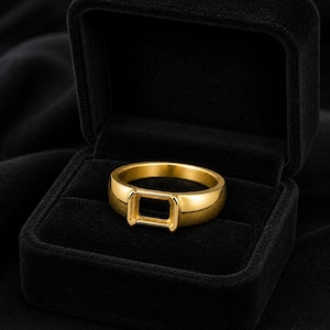 May include: A polished gold-tone ring featuring a smooth, rounded band and a prominent, rectangular-shaped setting at the center designed to hold a stone. The ring is displayed inside a black velvet-lined jewelry box with an open lid.