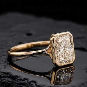 Könnte beinhalten: Ein Goldring mit einem rechteckigen Stein im Radiant-Schliff in einer glatten Zargenfassung. Die Ringschiene besteht aus poliertem Gelbgold mit einem schlanken Profil, das sich zur Fassung hin verjüngt und einen klaren, facettierten Edelstein zeigt.