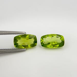 Könnte beinhalten: Zwei facettierte, rechteckige Peridot-Edelsteine mit einem leuchtenden, hellgrünen Farbton. Ein Edelstein wird von einer silbernen Pinzette gehalten. Die Edelsteine werden vor einem schlichten weißen Hintergrund präsentiert, der ihre Klarheit und Farbe hervorhebt. Ideal für die Schmuckherstellung.