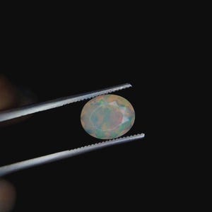 Può includere: Una gemma di opale ovale, tenuta da una pinzetta su sfondo nero. L'opale mostra un gioco di colori, con lampi iridescenti di verde, blu e rosa. La gemma è il punto focale, mostrando la sua bellezza naturale e i suoi intricati motivi.