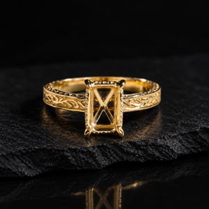 May include: A gold ring featuring an empty rectangular prong setting designed for a gemstone. The band displays an intricate, raised scrollwork pattern along its entire outer surface, providing a textured appearance to the polished metal finish.
