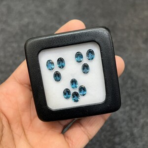 Könnte beinhalten: Eine schwarze quadratische Box mit weißem Innenraum enthält neun ovale, blaue Edelsteine. Die Edelsteine sind in drei Reihen mit jeweils drei Steinen angeordnet. Die Box wird in einer Hand gehalten.