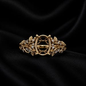 May include: A gold ring setting featuring an empty oval center mount designed for a gemstone. The band is crafted with intricate metalwork resembling branching vines and leaves, accented with small, clear, round-cut sparkling stones.