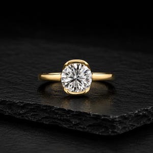 May include: A gold ring featuring a single round brilliant-cut clear gemstone set in a partial bezel setting. The band is smooth and polished, showcasing a high-shine metallic finish that contrasts with the faceted, sparkling white center stone.
