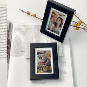 May include: Two black picture frames, one angled, displaying photographs. The top frame has a photo with the word "THOAT" visible. The frames rest on an open book with yellow decorative sprigs.