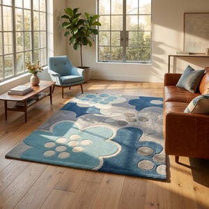 May include: A large area rug with a floral pattern in shades of blue, gray, and white. The rug features oversized flower shapes with a textured appearance. The rug is placed on a hardwood floor in a well-lit room with large windows.