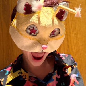 May include: A handmade cat mask with a yellow face, white ears, and red star accents. The mask has pink nose and whiskers. The person is wearing a black shirt with a colorful animal print.