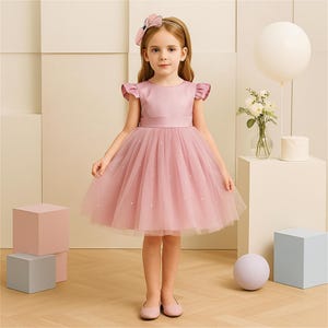 May include: A young girl wearing a pink dress with a tulle skirt and a matching headband. The dress has a satin bodice and a ribbon belt. The skirt is adorned with small, white, pearl-like embellishments. The girl is wearing pink shoes.