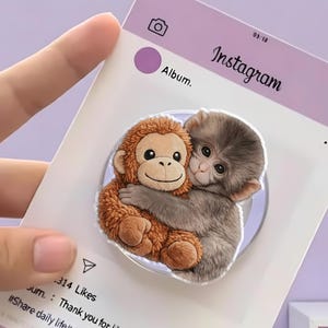 May include: A clear phone grip featuring a cartoon illustration of two hugging monkeys. One monkey is brown, the other is gray. The grip is on a white card with the Instagram logo and the word "Album."