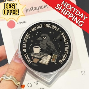 以下が含まれることがあります： カラスのイラスト、コーヒーカップ、そして「HIGHLY INTELLIGENT - MILDLY UNSTABLE - BARELY FUNCTIONAL」の文字が入った、白黒の丸いスマホグリップ。 グリップは手で持たれています。