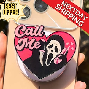 May include: A pink and black phone grip featuring a heart-shaped design with the text "Call Me". The design includes a ghost face with a knife and a phone. The phone grip is attached to a gold phone.
