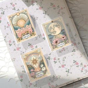 Peut inclure: Trois supports de téléphone avec des cartes de tarot représentant Le Soleil, La Lune et L'Étoile. Chaque carte présente une illustration unique aux couleurs pastel et des accents dorés. Les supports sont posés sur une surface à motifs floraux.