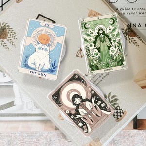 Puede incluir: Tres agarres para teléfono con cartas del tarot. Uno presenta un gato y el texto "THE SUN". Otro muestra a una mujer y el texto "EMPRESS". El tercero representa a una mujer con una luna creciente. Todos están sobre bases transparentes.