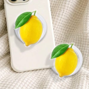 May include: A white phone case with a lemon-themed phone grip attached. The grip features a bright yellow lemon with a green leaf. A second lemon grip is visible next to the phone.