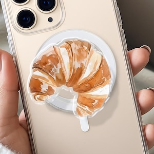 May include: A clear phone case with a watercolor-style croissant design on a white circular grip. The phone has a triple-lens camera. The case is held by a person wearing a white sweater.