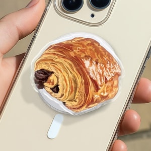 May include: A clear phone case with a detailed image of a chocolate croissant. The case is beige and has a clear border. The croissant is golden brown with dark chocolate filling. The phone has three camera lenses.