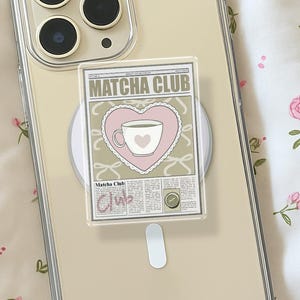 May include: A clear phone case with a Matcha Club graphic. The graphic features a pink heart with a teacup design and the words "Matcha Club". The phone case is on a floral patterned surface.