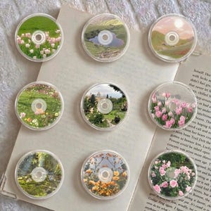 May include: Nine clear CD-shaped coasters with various landscape designs. Each coaster features a different miniature scene, including fields of flowers, a lake, and a garden. The coasters are arranged on a book page.