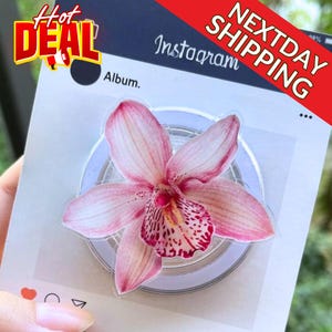 May include: A pink and white floral phone grip with a detailed orchid design. The grip is mounted on a white and silver base. The image also includes the text "Hot Deal", "Next Day Shipping", and "Instagram Album."