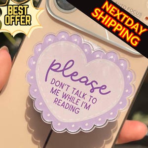 Może przedstawiać: Uchwyt do telefonu w kształcie serca z falistą krawędzią i napisem "please DON'T TALK TO ME WHILE I'M READING" w fioletowym skrypcie. Uchwyt jest jasnofioletowy i biały. Obraz zawiera również tekst "BEST OFFER" i "NEXTDAY SHIPPING".
