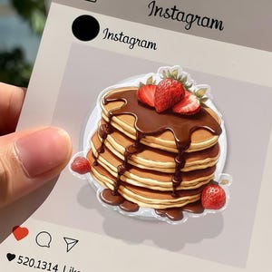 May include: A stack of pancakes with chocolate syrup and strawberries, in a sticker design. The sticker has an Instagram theme, with the word "Instagram" at the top. The pancakes are golden brown, and the strawberries are red.