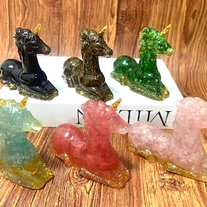 May include: Six resin unicorn figurines in various colors: black, brown, green, teal, red, and pink. Each has a golden horn and base, with glitter accents. The unicorns are displayed on a white surface.