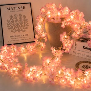 Peut inclure: Une guirlande de fleurs de cerisier artificielles intégrant des lumières LED blanc chaud. La guirlande florale rose est drapée autour d'un vase aux tons neutres, créant une lueur douce. Les pétales délicats sont illuminés par de petites lampes.