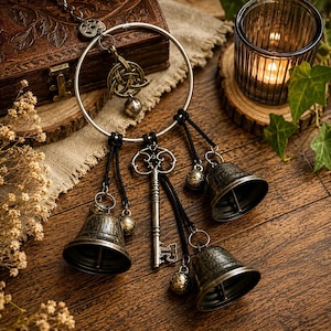 Può includere: Una decorazione da parete composta da un grande anello in metallo ornato con un ciondolo triquetra color bronzo, una chiave in stile antico e tre campanelli in metallo invecchiato sospesi da cordini neri, creando un aspetto rustico e stratificato.