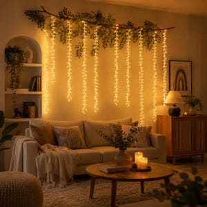May include: A horizontal tree branch mounted on a wall, adorned with green leaves and multiple vertical strands of warm white fairy lights that hang downward, creating a glowing curtain effect above a beige sofa and a wooden coffee table.
