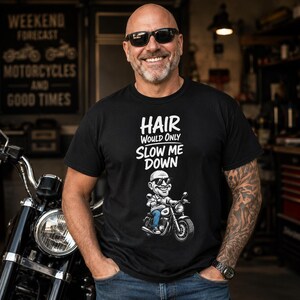 May include: A black short-sleeved t-shirt featuring white text that reads "HAIR WOULD ONLY SLOW ME DOWN" above a cartoon illustration of a bald person wearing sunglasses and riding a motorcycle while wearing blue jeans and a white shirt.